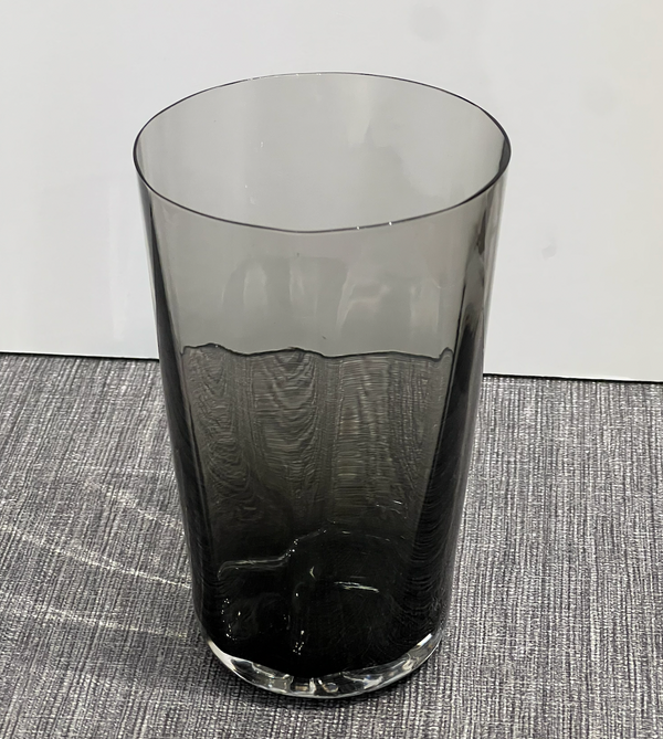 The Optic Cups Charcoal by Gary Bodker, a tall, cylindrical handcrafted glassware in translucent dark gray, sits on a textured gray surface against a plain white background.