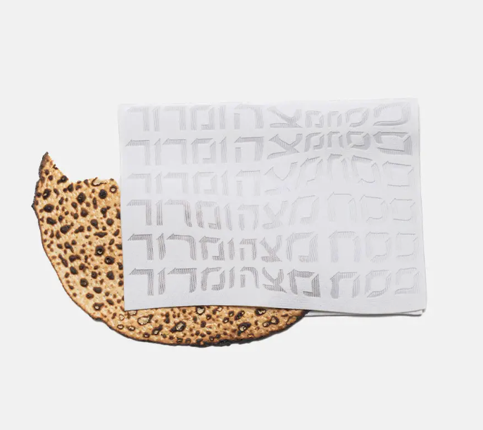 A piece of matzah rests on a white background beside the Apeloig Type Afikoman Bag, featuring a repeating laser-cut Hebrew design.