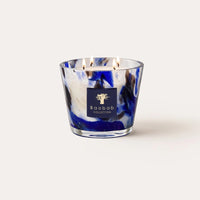 A lit Baobab Halona Candle Blue World, presented in a hand-blown glass holder with blue, black, and white swirls, sits against a plain light background. The woody fragrance fills the air with notes of rosemary, cedarwood, and vetiver for an inviting atmosphere.