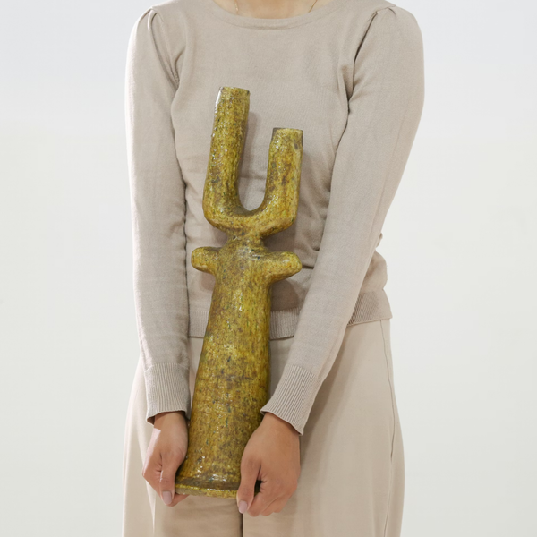 A person in a beige outfit holds the Govad Candleholder 20″ by Schwung Design—a large, gold, U-shaped sculpture with both hands. The candleholder’s two raised arms evoke the elegance of a classic candlestick and reflect the intricate essence of Moroccan artistry.