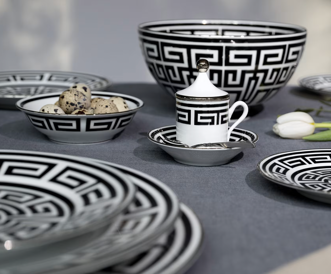 A table set with Ginori 1735’s Labirinto Dinnerware Nero—black and white geometric plates, bowls, and a lidded cup—plus quail eggs and a white tulip on gray linen exudes refined elegance.