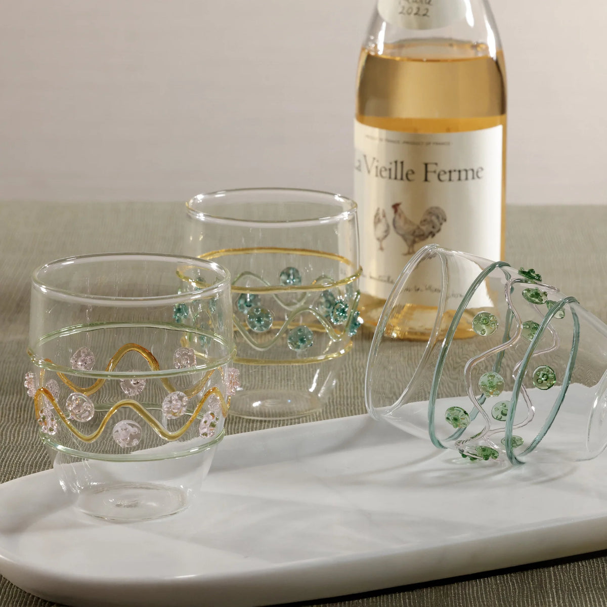 Three Zodax Festa Tumblers in green, amber, and pink tones are displayed on a white tray, with a bottle of La Vieille Ferme white wine in the background atop a neutral tabletop.