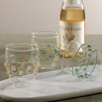 Three Zodax Festa Tumblers in green, amber, and pink tones are displayed on a white tray, with a bottle of La Vieille Ferme white wine in the background atop a neutral tabletop.