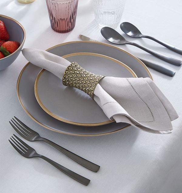 A neatly set table features two gray plates, a bowl of strawberries, cutlery, and a Sferra Festival Napkin (set of 4) in a gold-patterned ring on a white tablecloth, with two drinking glasses placed above the setting.
