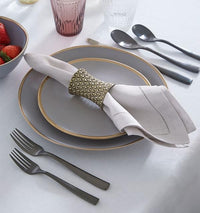 A neatly set table features two gray plates, a bowl of strawberries, cutlery, and a Sferra Festival Napkin (set of 4) in a gold-patterned ring on a white tablecloth, with two drinking glasses placed above the setting.
