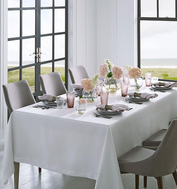 A dining table set for six with a white tablecloth, grey chairs, Sferra Festival Napkin Set of 4 in European linen, pink and clear glassware, pink floral centerpieces, and large windows overlooking grass and water.