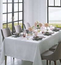 A dining table set for six with a white tablecloth, grey chairs, Sferra Festival Napkin Set of 4 in European linen, pink and clear glassware, pink floral centerpieces, and large windows overlooking grass and water.