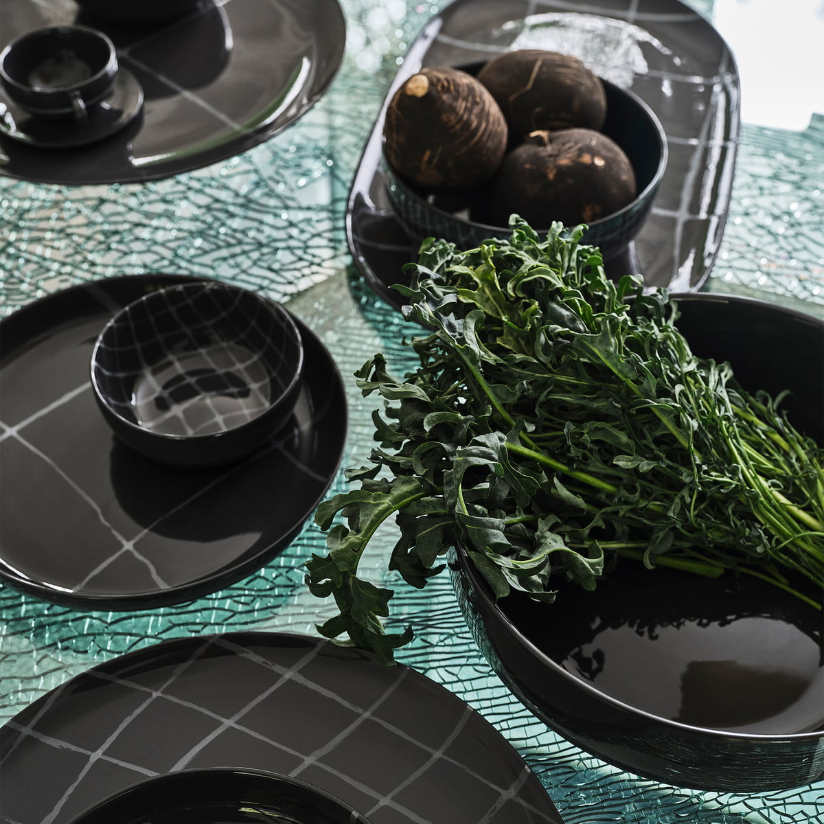 A table set with Serax Zuma Dinnerware Pacific—porcelain plates, bowls, and serving dishes with Bauhaus-inspired grids—holds leafy greens and root vegetables on a textured glass surface.