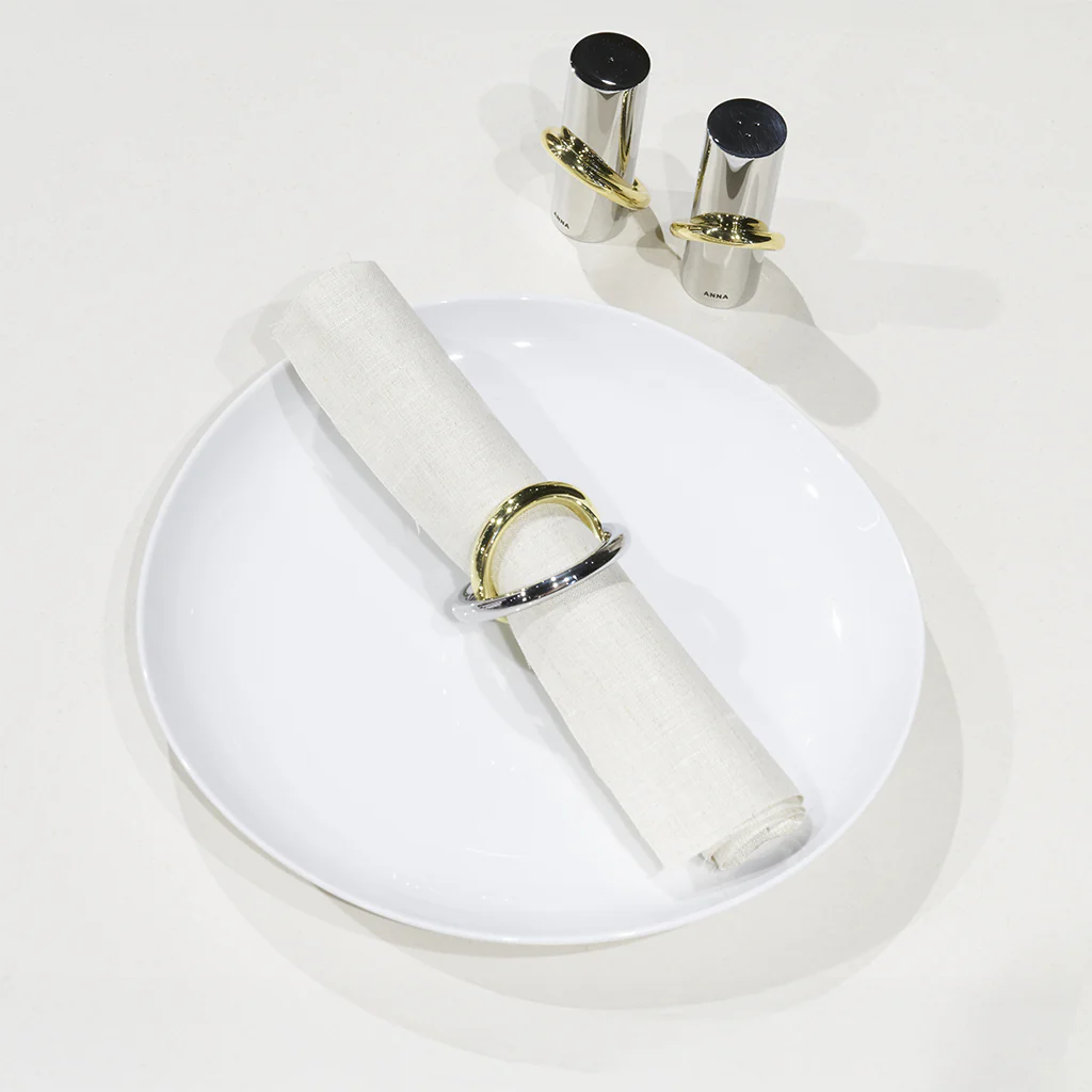 A white plate holds a neatly rolled white napkin with a brass and stainless steel napkin ring. In the background, the Anna New York Ring Salt & Pepper Shaker Set in silver and gold stands on a white surface.