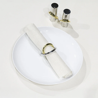 A white plate holds a neatly rolled white napkin with a brass and stainless steel napkin ring. In the background, the Anna New York Ring Salt & Pepper Shaker Set in silver and gold stands on a white surface.