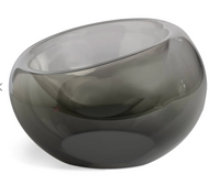 The Axiom Glass Caldera Vessel Medium 9" is a round, transparent gray hand-blown glass piece with a smooth, glossy finish and an asymmetrical tilted rim, shown against a white background.
