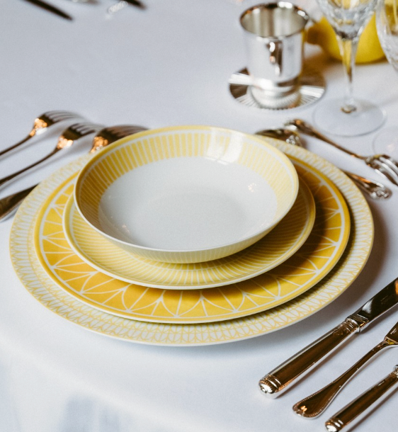 A close-up of a table setting with Christofle’s Malmaison Riviera Dinnerware, silver cutlery, a wine glass, and a silver cup, all arranged on a white tablecloth evoking French Riviera dining.