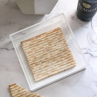 A single matzah rests in the BT-Shalom Lucite Matzah Box With Lid on a clear tray over marble, with another matzah and a wine bottle nearby.