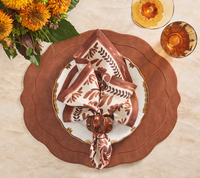 A cozy autumn table is set with brown and white plates, amber glasses, orange flowers, and Kim Seybert's Botanica Napkin Ivory & Brown (Set of 4), featuring leaf patterns and a pumpkin-shaped ring on a beige surface.