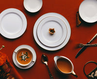 A refined table features Bernardaud's Palmyre Dinnerware—white plates with gold rims—set on a red tablecloth, complemented by gold cutlery, a bowl of soup garnished elegantly, and a small pastry for an understated yet elegant look.