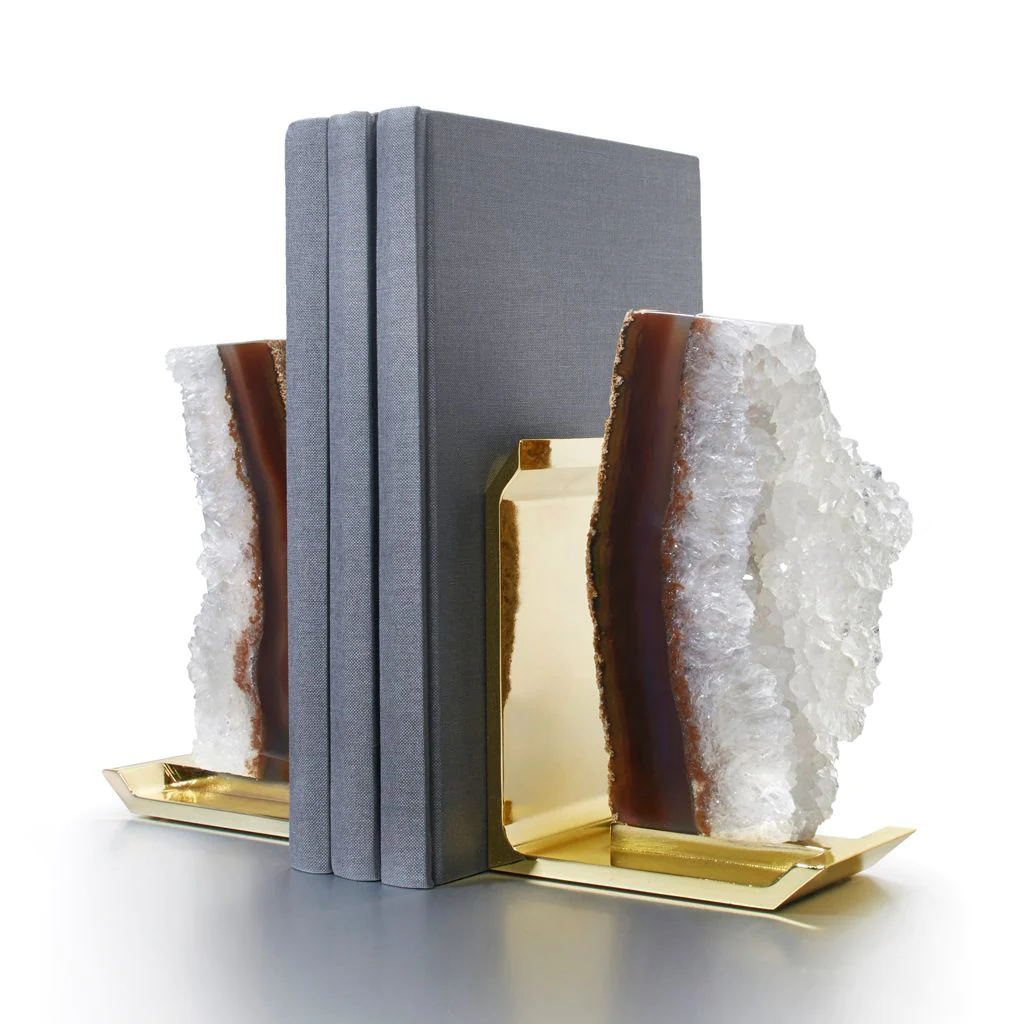Two gray hardcover books stand upright between the Fim Agate Druze & Gold Bookend Set by Anna New York, featuring large, rough-edged crystals with a sparkly white and brown geode look, set against a plain white background.