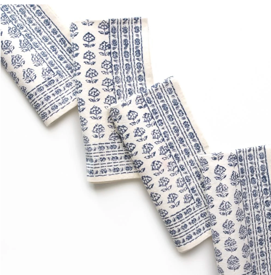 The Elise Napkin Set of 4 by Pomegranate features a blue floral and geometric pattern on white, displayed in a zigzag layout beside layered blue tableware on a white surface.