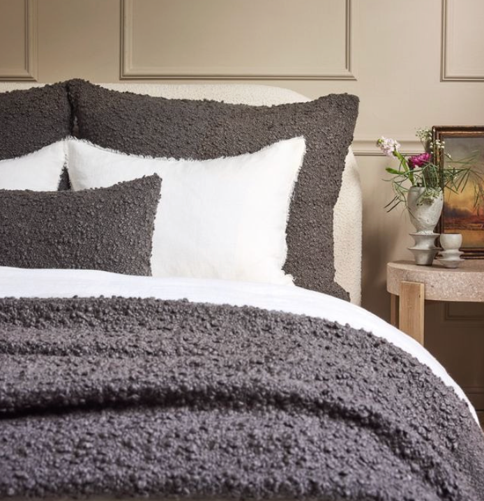 A neatly made bed features the Murphy Throw - Midnight by Pom Pom At Home, styled with textured dark gray and white pillows. Beside it sits a small table holding a glass vase of flowers, a cup and saucer, and a framed painting.