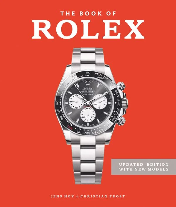 A silver Rolex watch with a black and silver face sits centered on the red cover of The Book of Rolex by ACC ART BOOKS / SIMON & SCHUSTER—an updated guide to spotting fake Rolex watches and discovering new models.