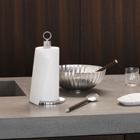 A paper towel roll on a metal holder, a silver bowl with chopsticks, a glass, the Georg Jensen Bernadotte Stainless and Wood Salad Set spoon, and a small dish are arranged on a modern gray kitchen countertop. Dark wooden cabinets are in the background.