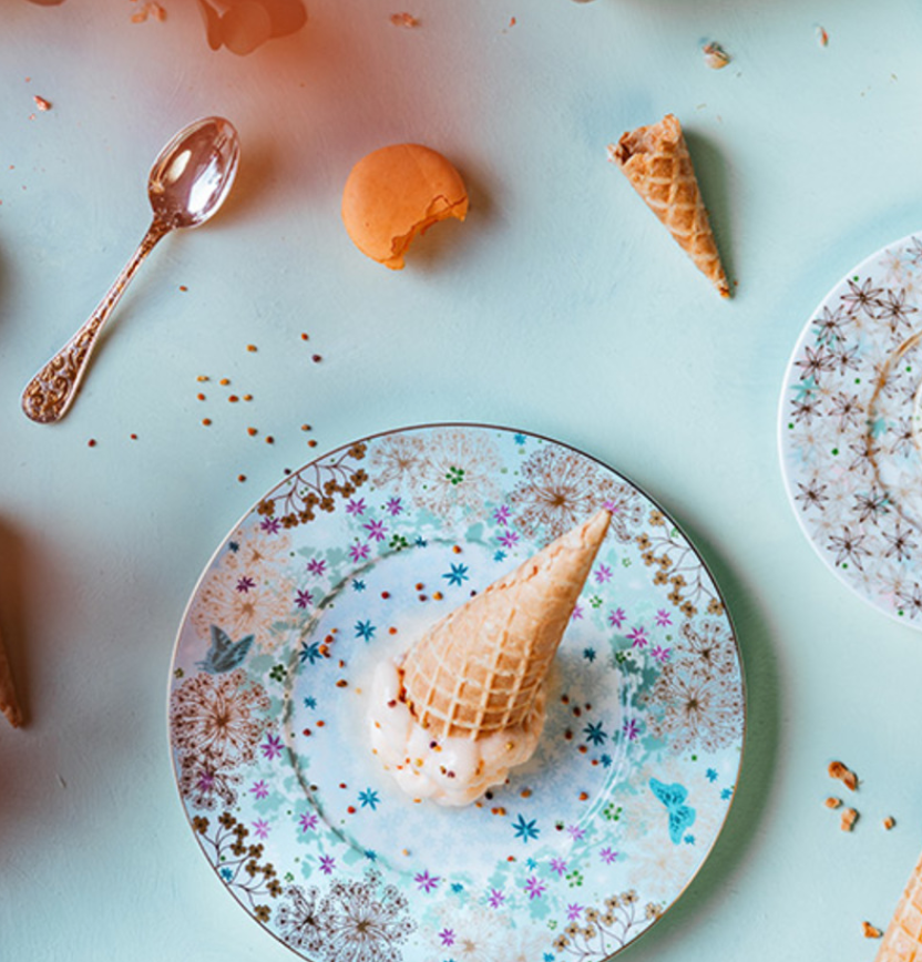 The Fèerie By Michael Cailloux plate by Bernardaud features an upside-down melting ice cream cone with crumbs, a broken wafer, and a spoon beside another decorated plate on a light blue surface.