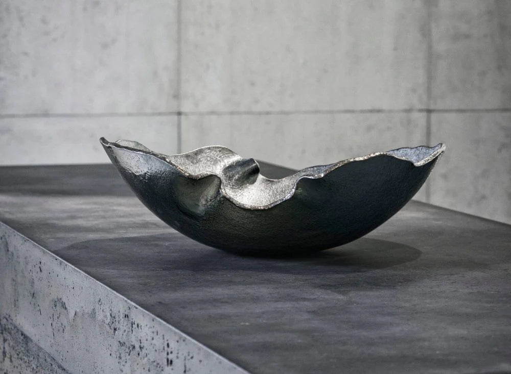 The Gardeco Mapa Grey & Silver Trim Bowl, featuring a dark, metallic finish and wavy edges, rests on a textured gray concrete surface with a light gray concrete wall in the background.