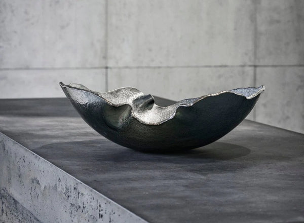 The Gardeco Mapa Grey & Silver Trim Bowl, featuring a dark, metallic finish and wavy edges, rests on a textured gray concrete surface with a light gray concrete wall in the background.
