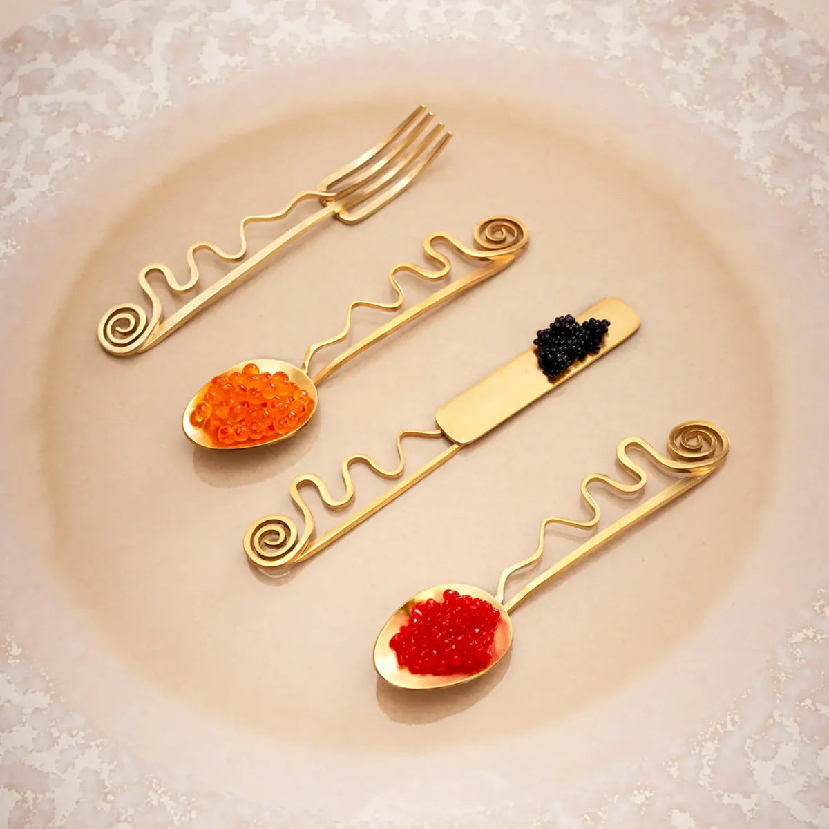 Four Spirale Gold Cocktail Forks by L'Objet, plated in 24k gold, are elegantly arranged on a decorative plate—each adorned with red, orange, or black caviar. Ideal for luxury presentation in a premium gift box.