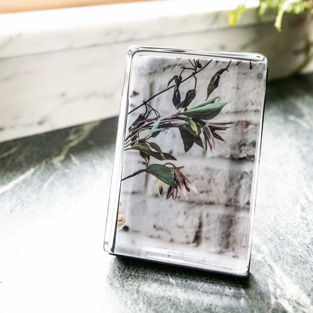 The Simon Pearce Woodbury Photo Block Vertical displays a botanical image of green leaves and flowers, handcrafted in glass and set on a dark marble surface, softly lit by window light.