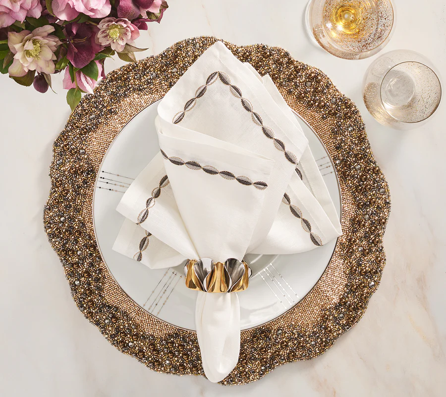 A decorative dinner setting features a gold beaded charger, white plate, embroidered napkin in a Kim Seybert Enigma Napkin Ring (Gold & Silver Set of 4), champagne glass, purple flower bouquet, and elegant gold and silver decor on white marble.