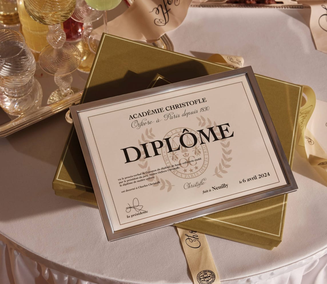 A Christofle Uni Picture Frame holds a French diploma dated April 6, 2024, atop a green box on a round table with glassware, ribbon, and decor—adding an elegant touch.