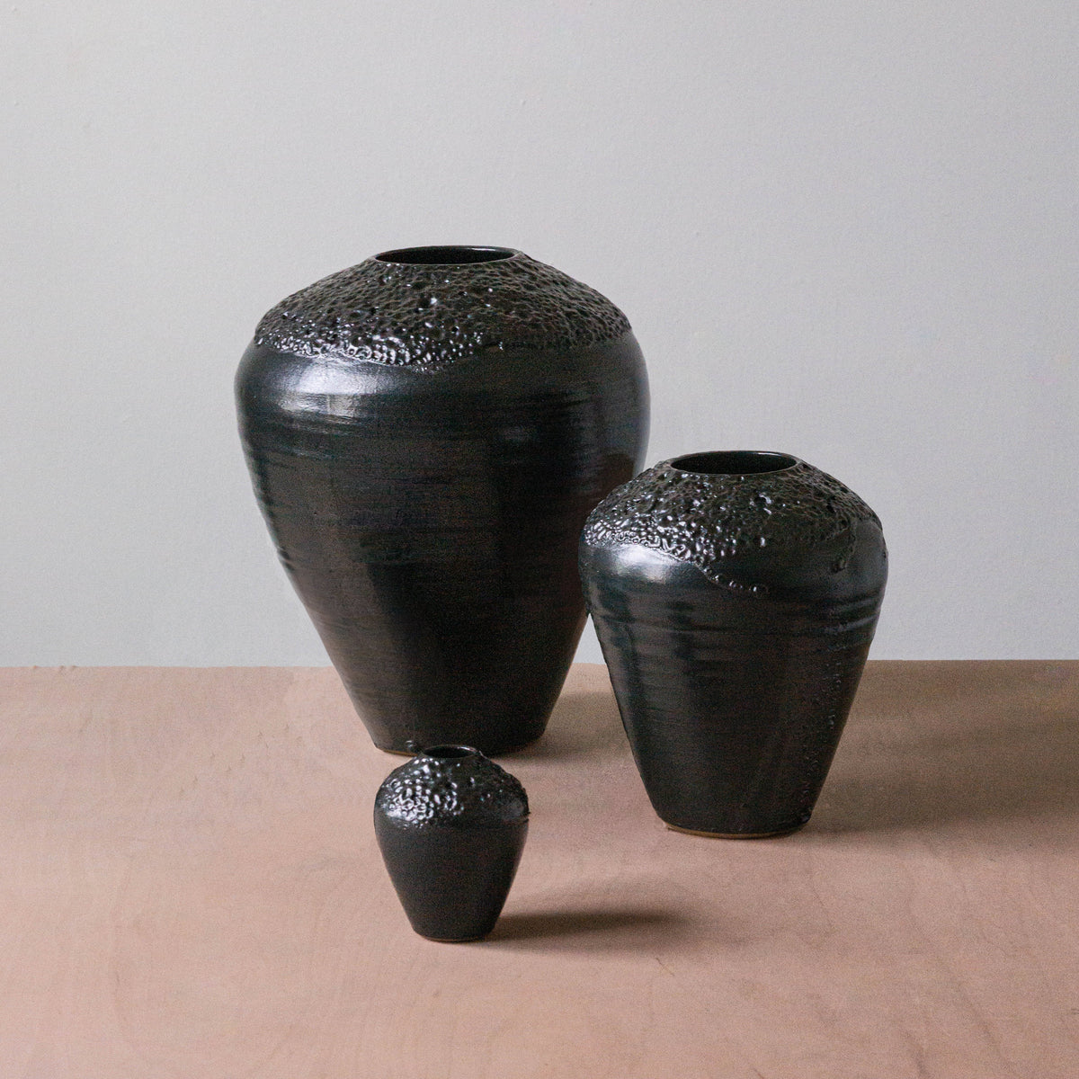 The Esme Crawl Vase - Black Lava by Kalya OD Studio features large and medium water-safe vases with black textured finishes, perfect for fresh or dried arrangements on any light brown surface against a gray wall.