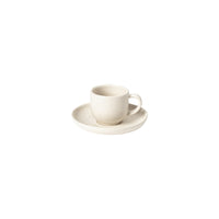 A small, white ceramic cup with a handle rests on a matching saucer from the Costa Nova Pacifica Vanilla Dinnerware collection. Both pieces feature a minimalist, slightly speckled finish and are set against a plain white background.