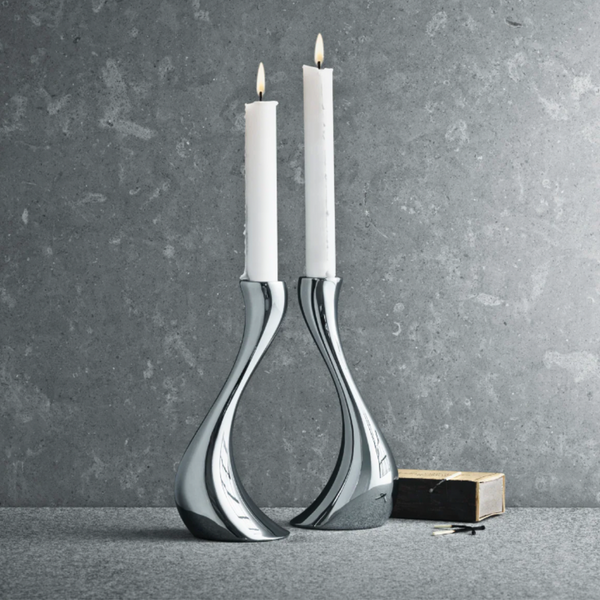 Two tall white candles glow in the Georg Jensen Cobra Candleholder 2 Piece Set, displayed on a gray stone surface with a matchbox and matches—ideal for adding a modern touch to minimalist home decor.