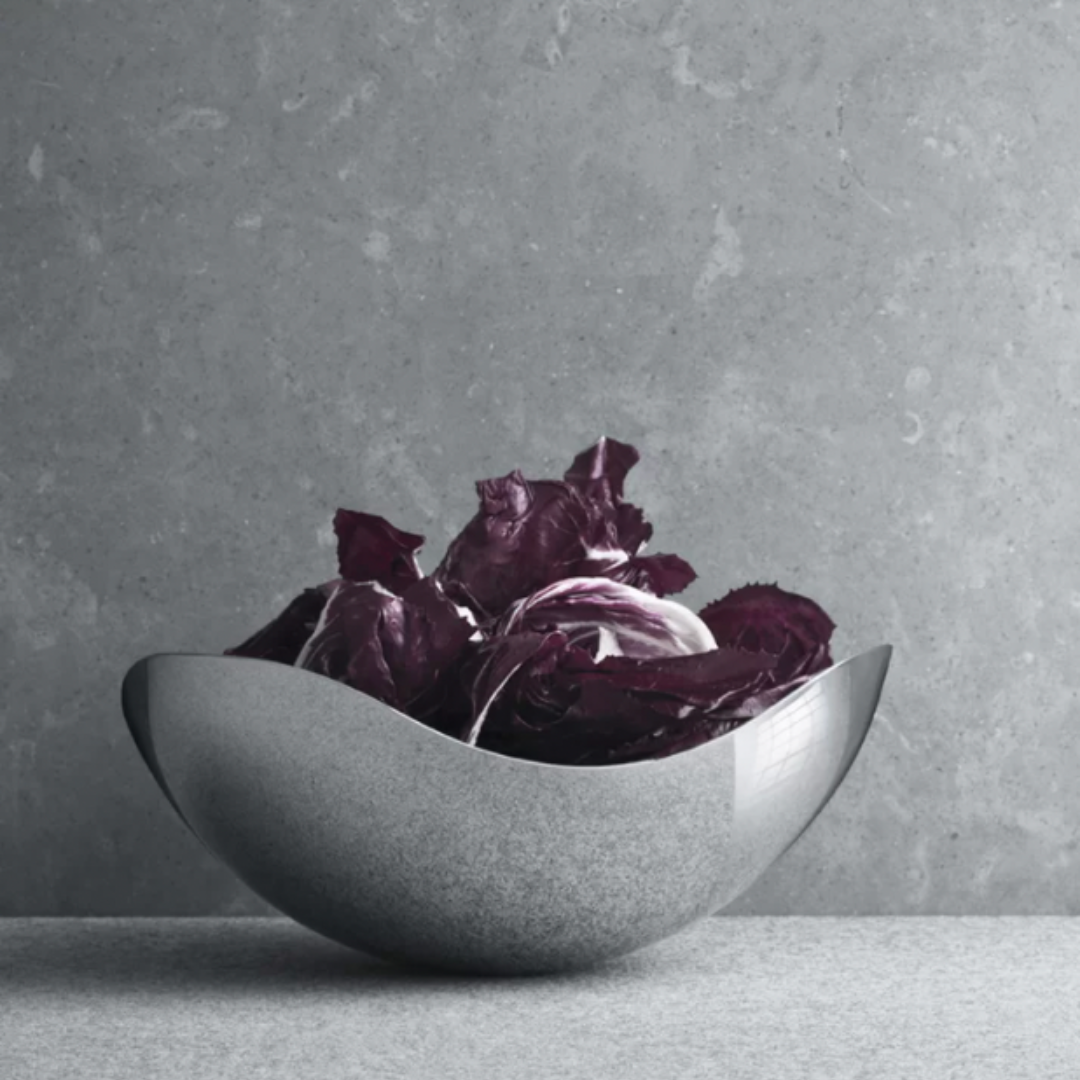 The Bloom Serving Bowl by Georg Jensen, with its modern curved stainless steel design and mirror-polished finish, elegantly cradles deep purple radicchio on a gray surface set against a textured gray backdrop.