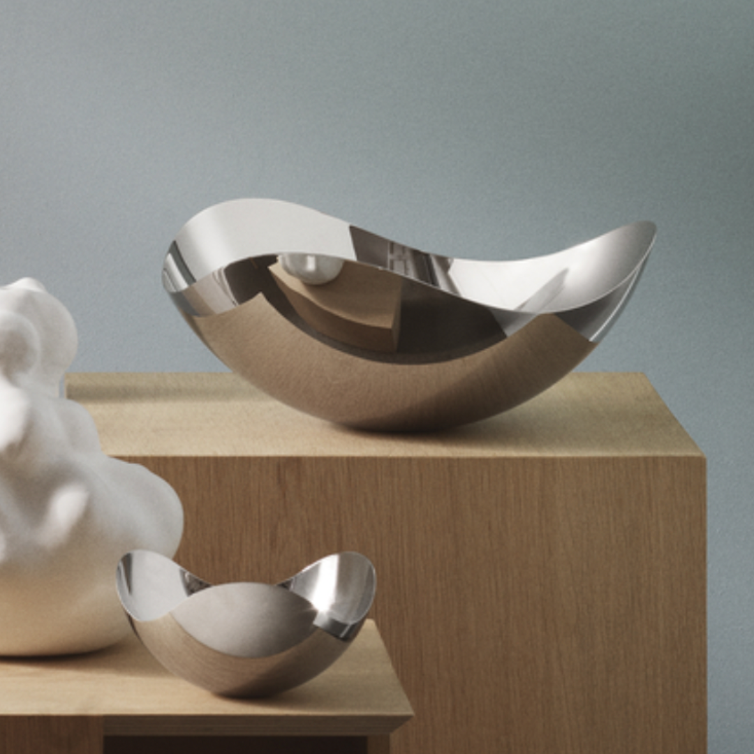 Two Georg Jensen Bloom Serving Bowls, crafted from mirror-polished stainless steel with curved edges, are set on wooden blocks beside a white abstract sculpture against a minimalist grey background.