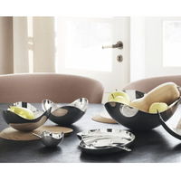 The modern dining table set highlights the Georg Jensen Bloom Serving Bowl with a mirror-polished finish, curved stainless steel design containing fruit, alongside a matching metal plate, spoon, and pale chairs near sunlit windows.