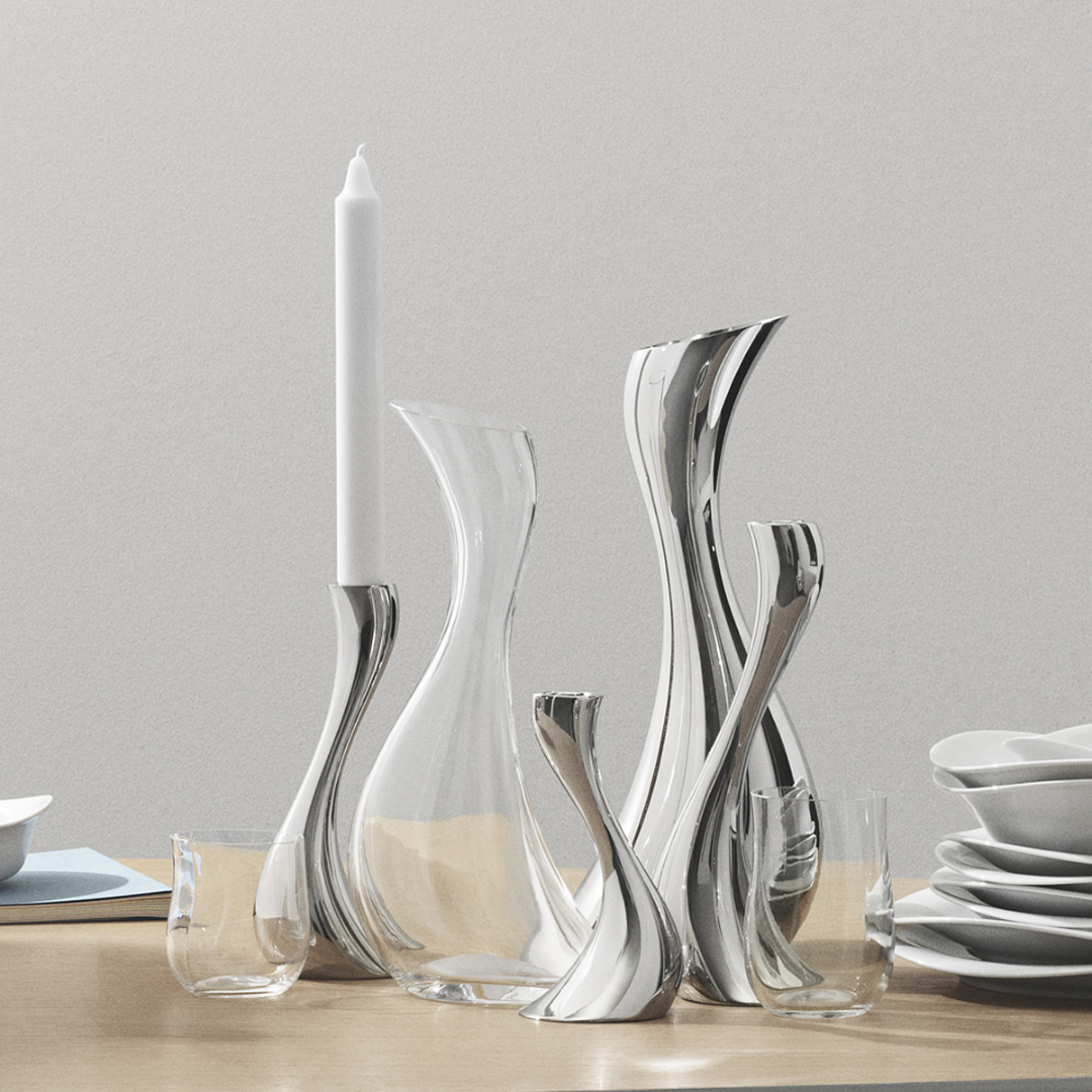 Elegant Georg Jensen metal and clear glass vases and candle holders with modern, organic shapes stand on a wooden table beside stacked white dishes, a white candle, and the Cobra Carafe.