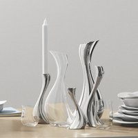 Elegant Georg Jensen metal and clear glass vases and candle holders with modern, organic shapes stand on a wooden table beside stacked white dishes, a white candle, and the Cobra Carafe.