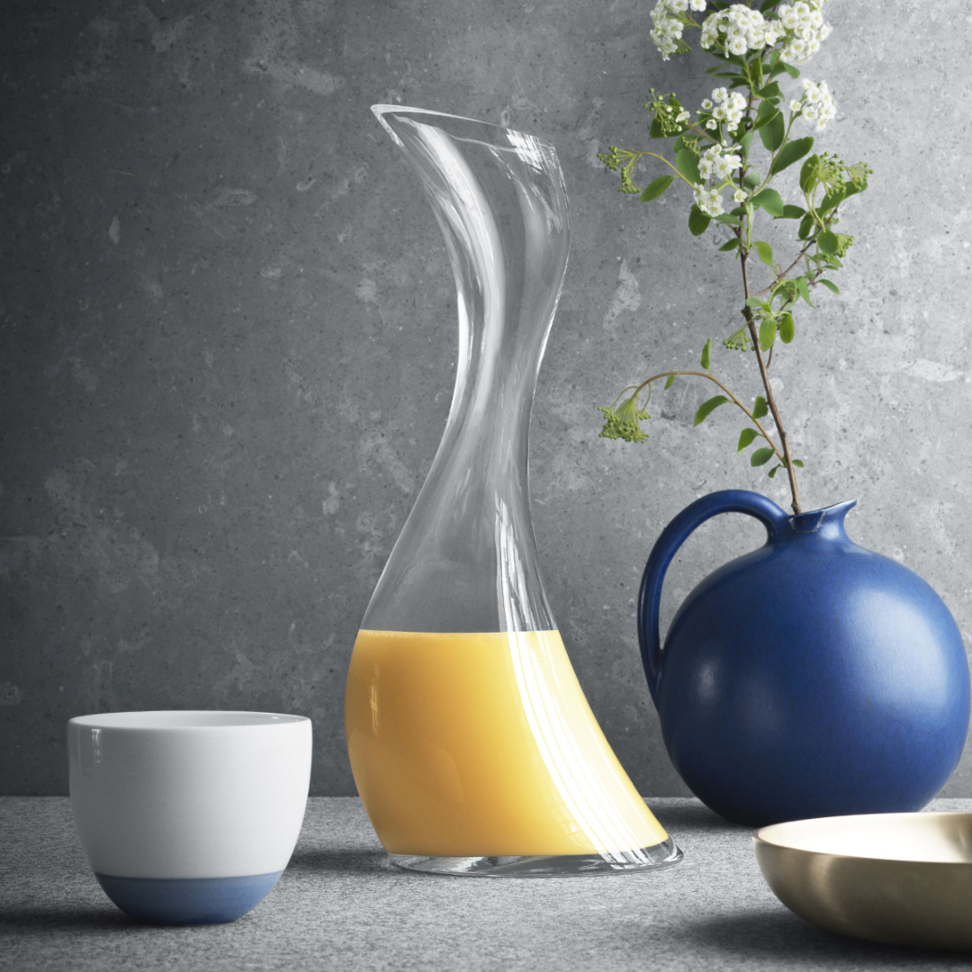The Georg Jensen Cobra Carafe, with its organic clear glass design, holds orange juice on a grey surface beside a blue ceramic jug of white flowers, a matching white-and-blue cup, and a cream-colored dish.
