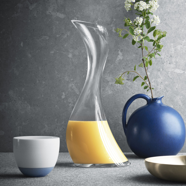 The Georg Jensen Cobra Carafe, with its organic clear glass design, holds orange juice on a grey surface beside a blue ceramic jug of white flowers, a matching white-and-blue cup, and a cream-colored dish.