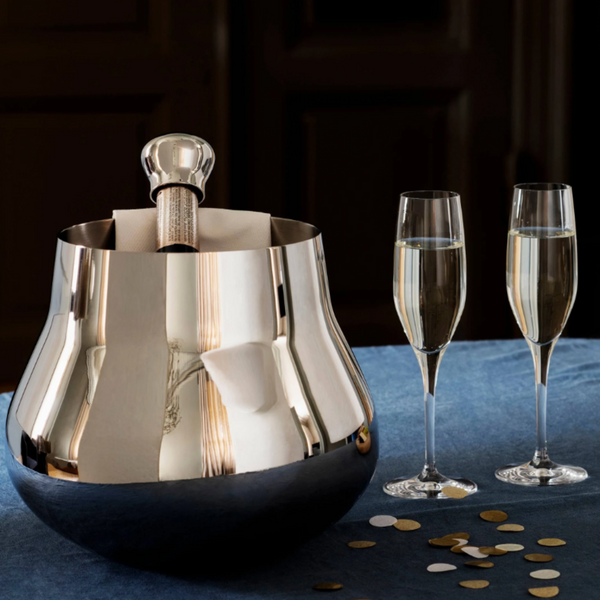 The Georg Jensen Sky Champagne & Wine Cooler, holding a bottle of champagne and napkin, rests on a blue tablecloth beside two filled flutes and scattered gold and white confetti.