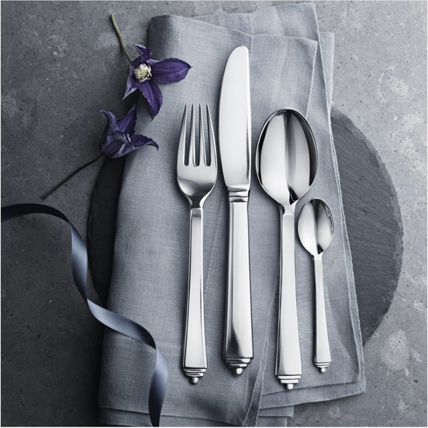 The Georg Jensen Pyramid Stainless Steel Mirror 5 Piece Flatware set is displayed on a folded gray napkin with a purple flower and ribbon, arranged on a gray stone surface.