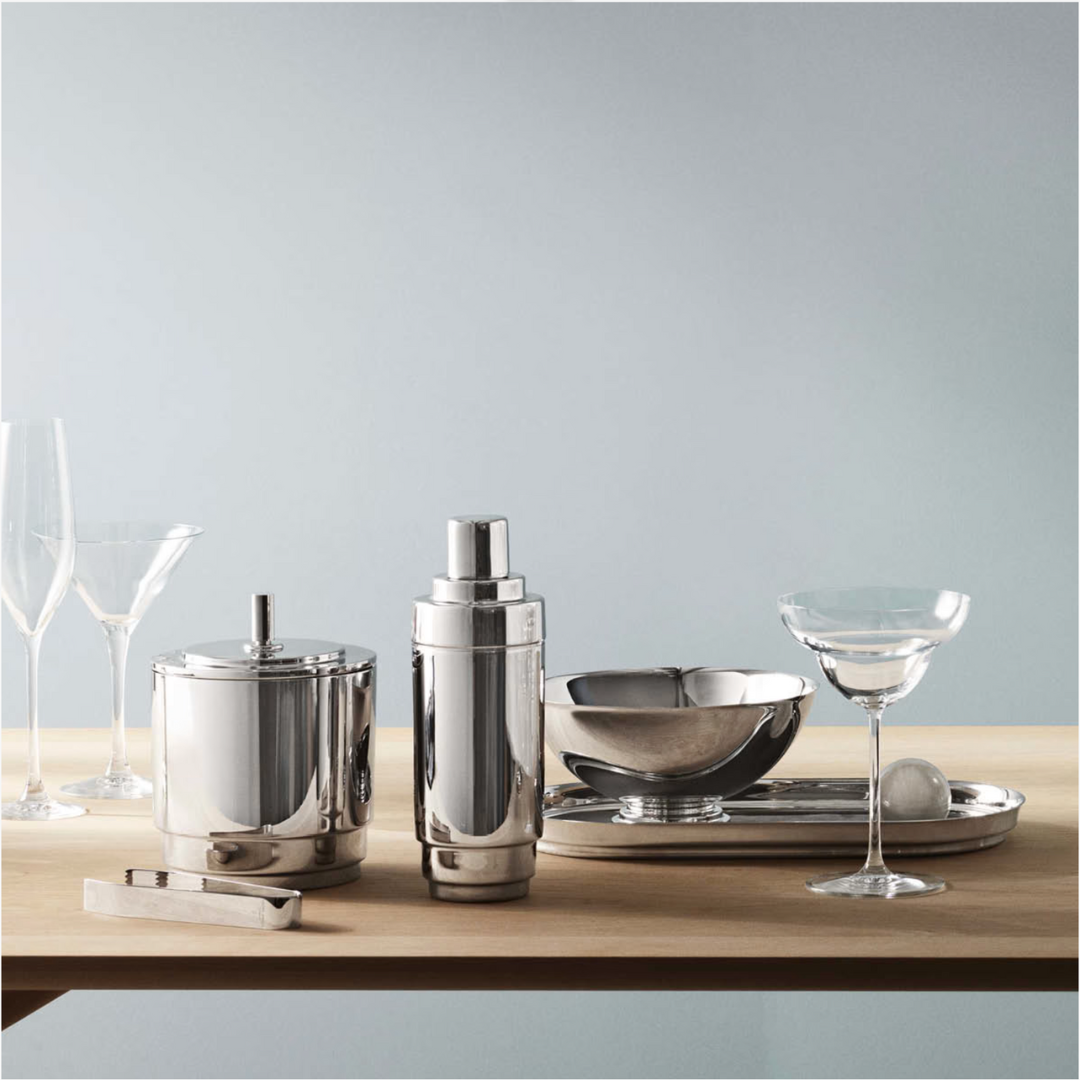 A Georg Jensen Manhattan Ice Bucket & Tong Set shines alongside a cocktail shaker, tray, bowl, and two glasses on a light wooden surface with a pale blue backdrop.