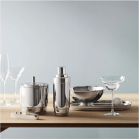 A Georg Jensen Manhattan Ice Bucket & Tong Set shines alongside a cocktail shaker, tray, bowl, and two glasses on a light wooden surface with a pale blue backdrop.