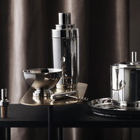 The Georg Jensen Manhattan Cocktail Shaker and matching barware—bowl, corkscrew, and ice bucket—are displayed on a black surface with a silky dark curtain behind, ideal for elevating your home bar.