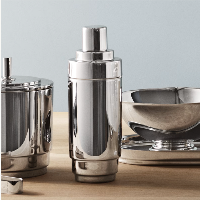 The Georg Jensen Manhattan Cocktail Shaker stands on a light wooden surface, accompanied by a matching ice bucket and metal bowl—a stylish upgrade for any home bar, set against a pale blue background.