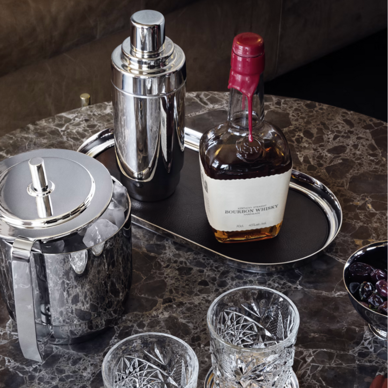 A marble table set with a Georg Jensen Manhattan Tray With Leather Liner, a silver ice bucket, cocktail shaker, bourbon whiskey bottle, two cut crystal glasses, and a bowl of dark cherries.