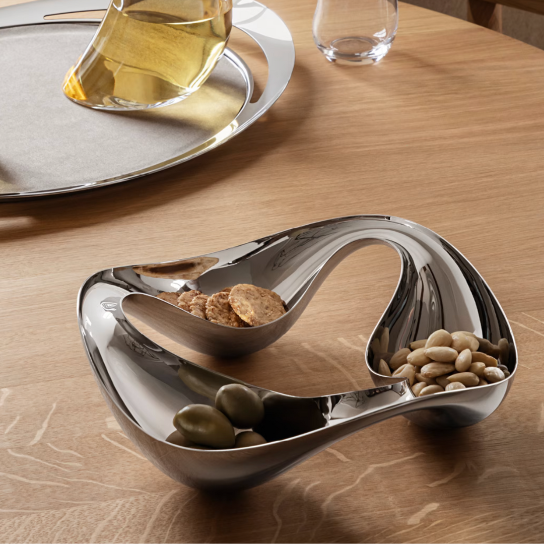 The Georg Jensen Cobra Snack Bowl, a modern sculptural stainless steel piece with three compartments, elegantly holds snacks on a wooden table; a glass decanter and tray in the background make it a striking centerpiece.