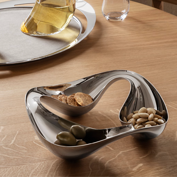 The Georg Jensen Cobra Snack Bowl, a modern sculptural stainless steel piece with three compartments, elegantly holds snacks on a wooden table; a glass decanter and tray in the background make it a striking centerpiece.